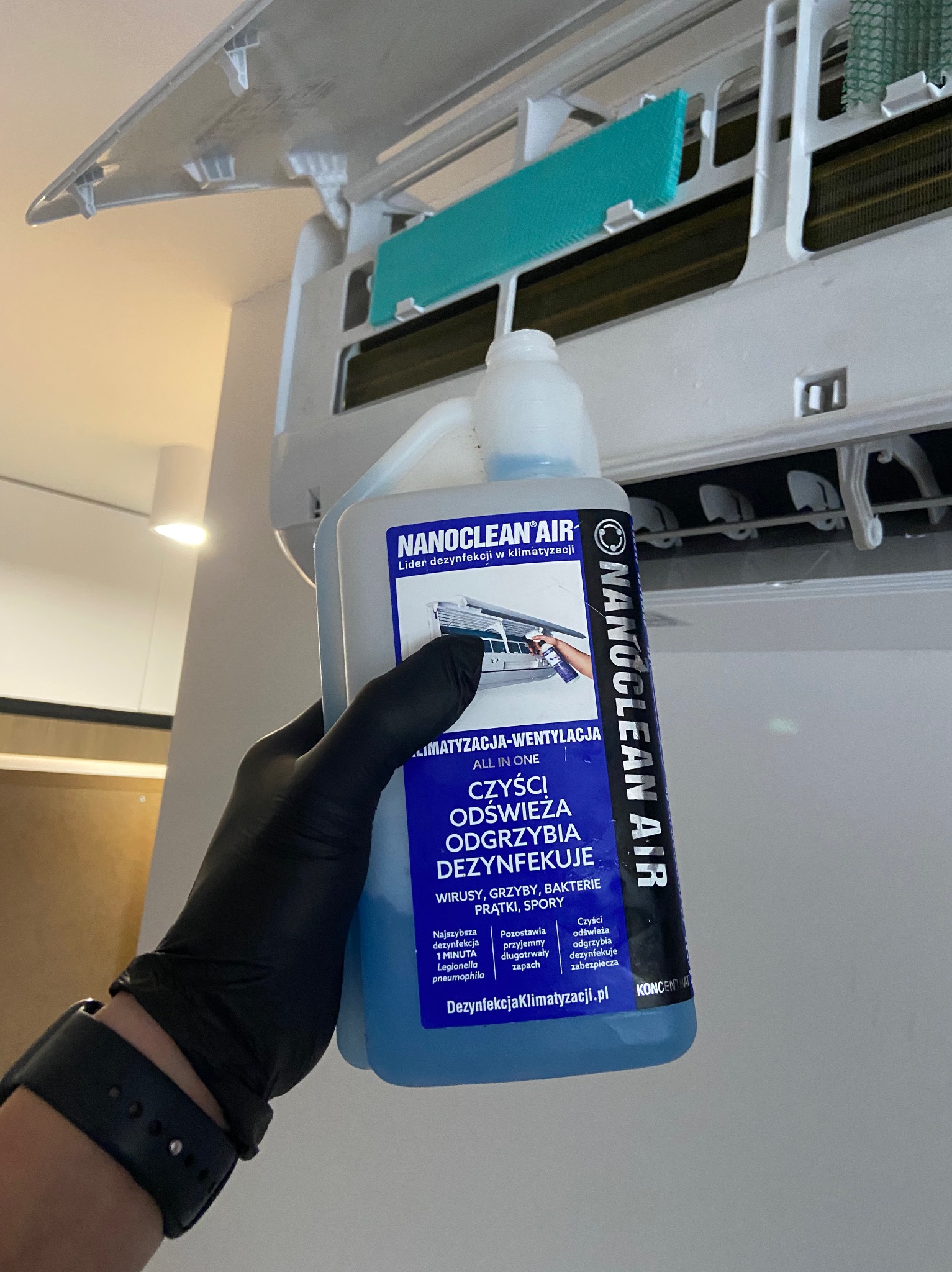 Czyszczenie klimatyzacji: dłoń w rękawicy trzyma butelkę środka dezynfekującego Nanoclean Air przed otwartym klimatyzatorem. Widoczne filtry i wnętrze urządzenia.