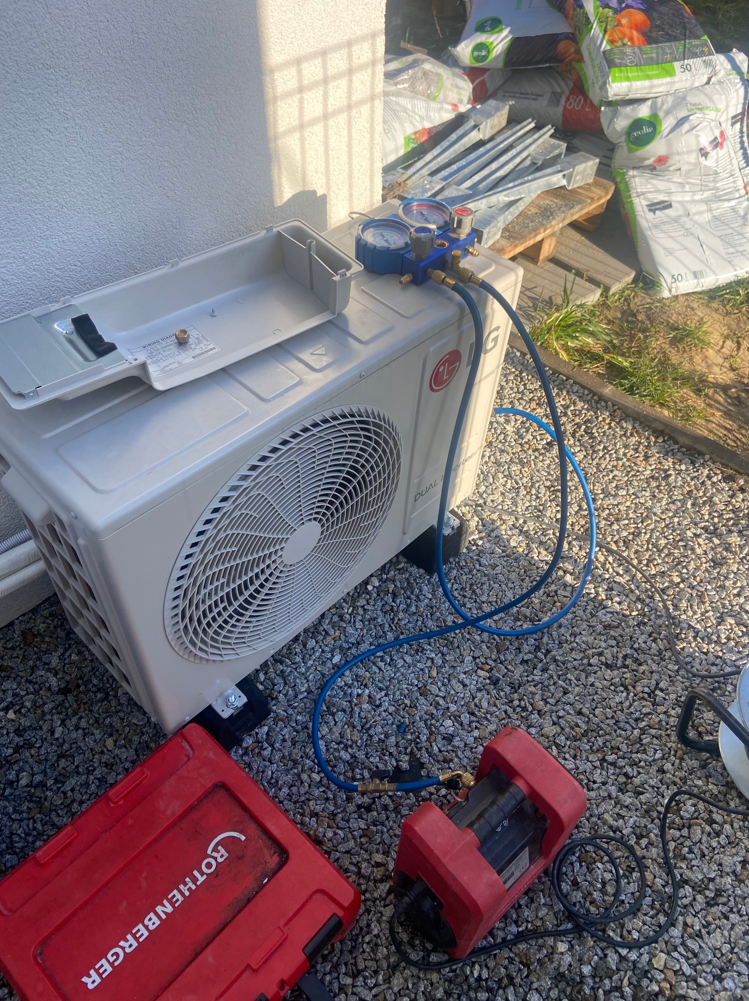 Biała jednostka zewnętrzna klimatyzacji LG, podłączona do manometrów i pompy próżniowej Rothenberger, na tle szarego żwiru i worków ziemi.