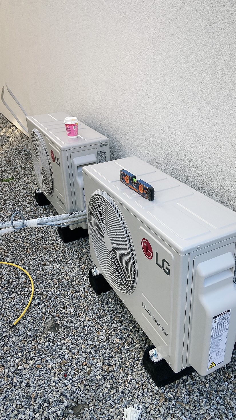 Dwa zewnętrzne moduły klimatyzacji LG Dual Inverter na żwirowym podłożu. Na jednym poziomica, na drugim kubek. Widoczne przewody i mocowania.