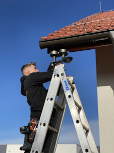 Mężczyzna na drabinie montuje kamerę pod okapem dachu; widoczne narzędzia w pasie, błękitne niebo w tle.