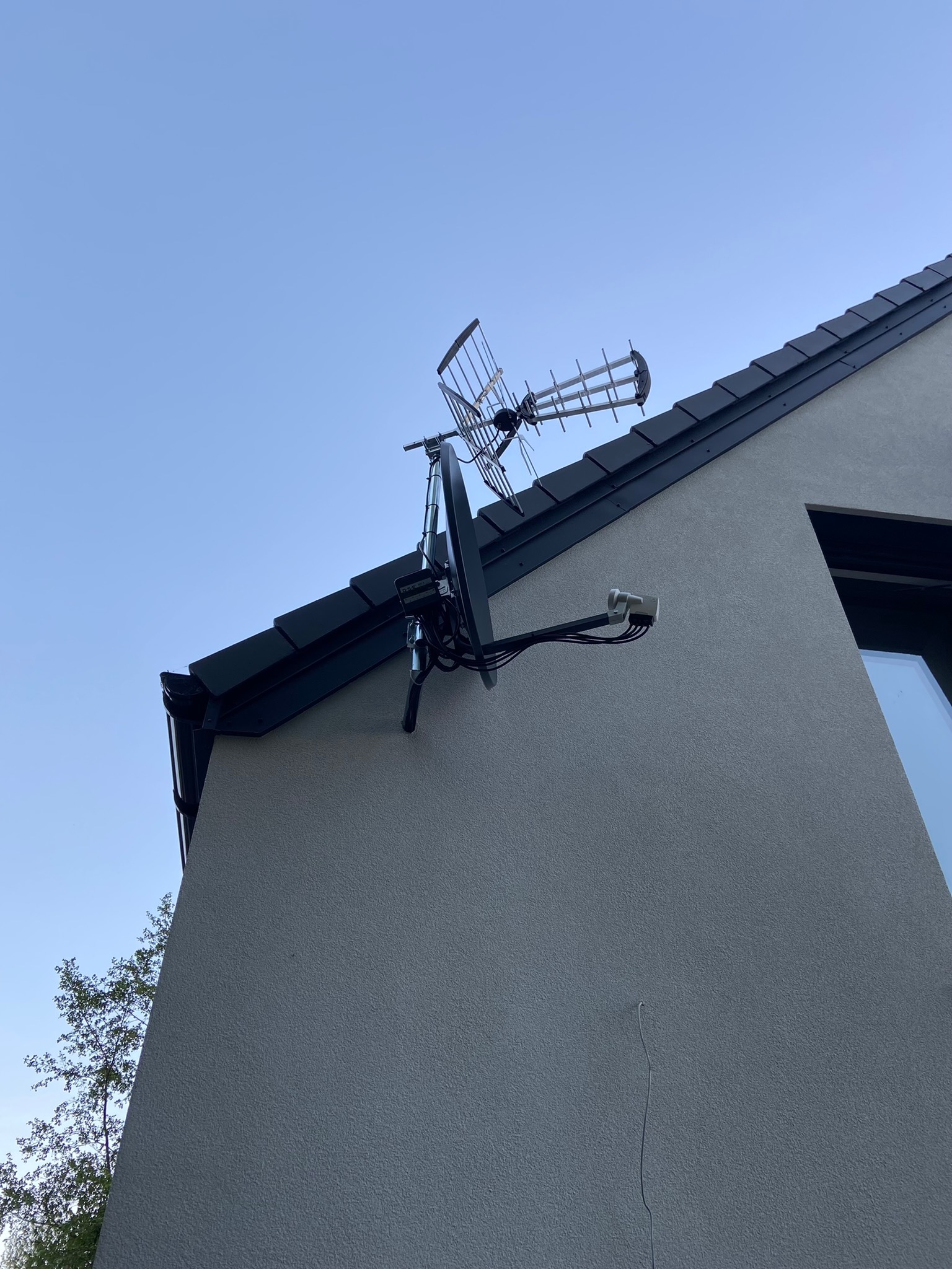 Antena telewizyjna zamontowana na elewacji domu jednorodzinnego, widok z dołu na tle jasnego nieba.