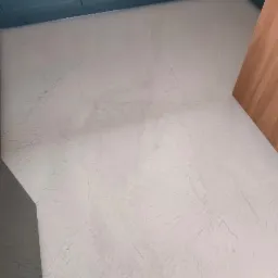 Jasnoszara podłoga z efektem chmurkowym, imitującym kamień, w rogu pomieszczenia z niebieskimi szafkami i drewnianym elementem wykończeniowym.