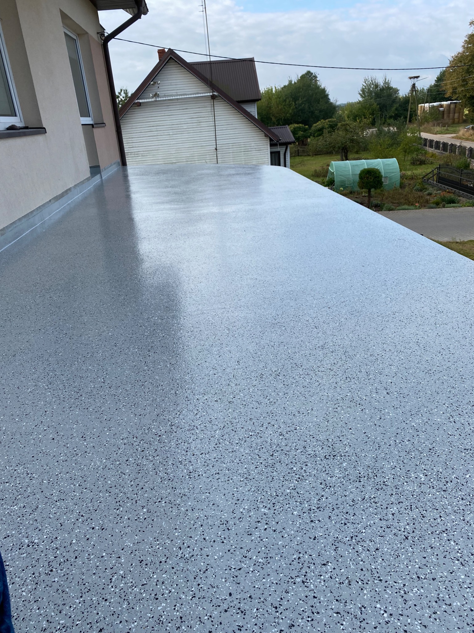 Szara, gładka powierzchnia balkonu z drobnymi, czarnymi i białymi elementami dekoracyjnymi, widok z góry na tle budynku mieszkalnego i zieleni.