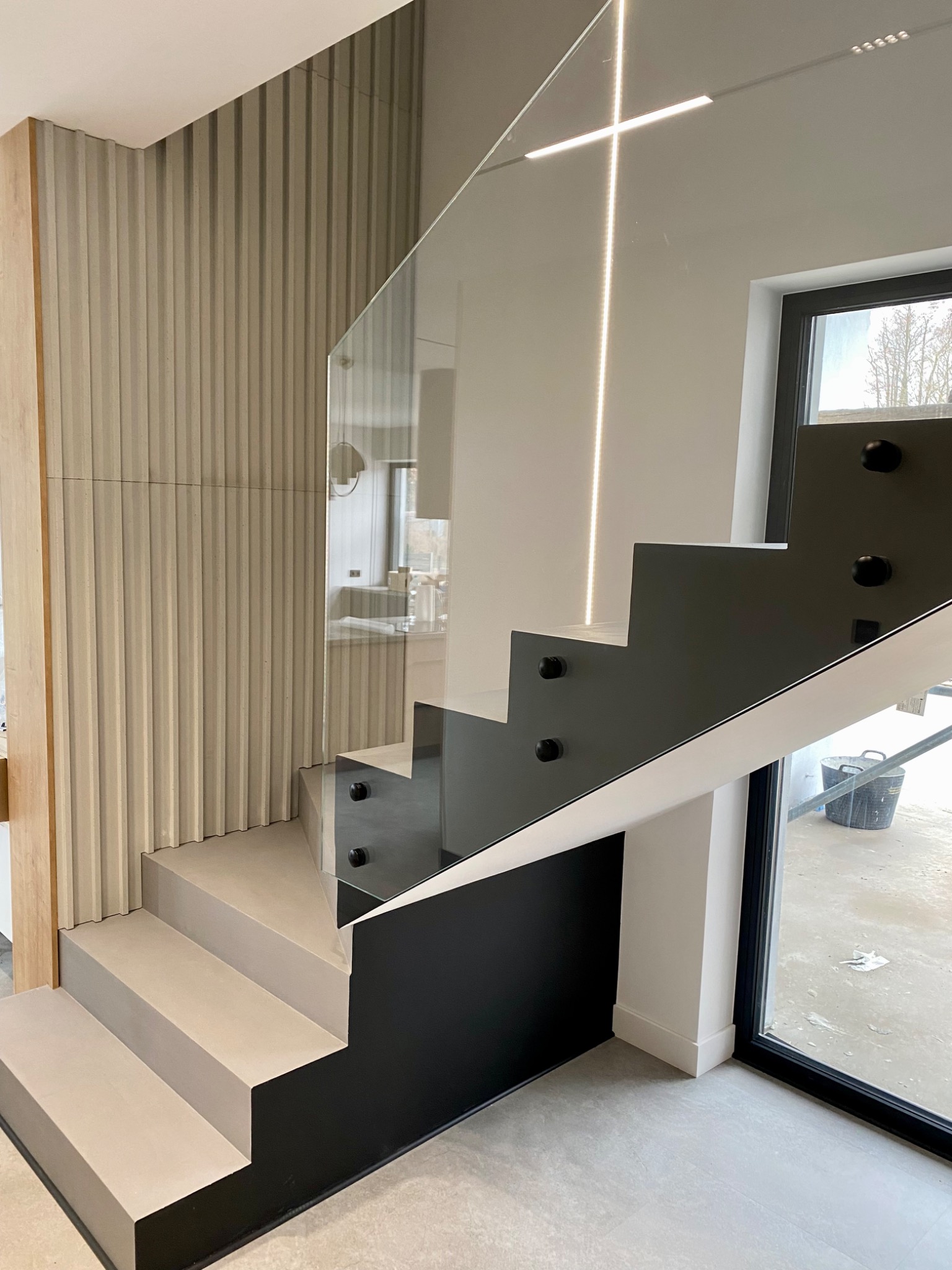 Nowoczesne schody z szarą, gładką powierzchnią stopni, szklaną balustradą z pionowym oświetleniem LED, czarnymi elementami konstrukcyjnymi i ryflowaną ścianą w tle.