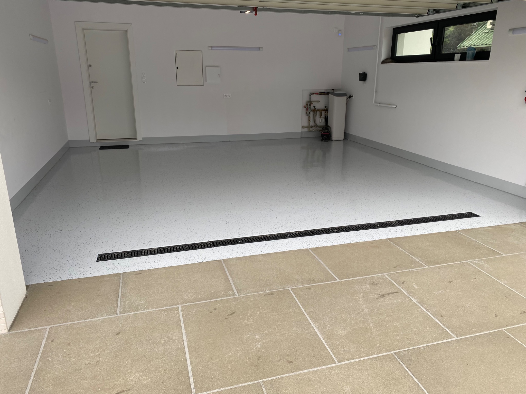 Jasnoszara, bezspoinowa posadzka z drobnymi, kolorowymi wtrąceniami w garażu, oddzielona czarnym odpływem liniowym od podjazdu wyłożonego kamiennymi płytami. Widoczna instalacja wodna na ścianie.