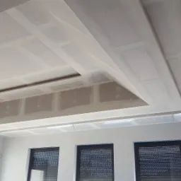 Sufit podwieszany z płyt kartonowo-gipsowych, z wnęką i oświetleniem w trakcie montażu, widoczne łączenia płyt i szpachlowanie, trzy okna z roletami w tle.