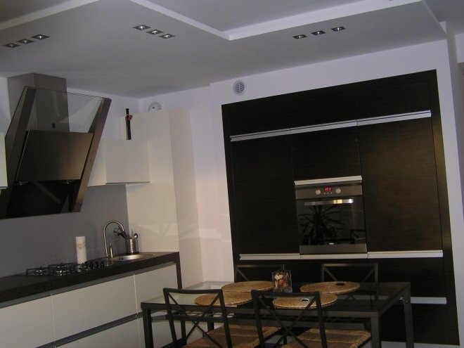 Nowoczesna kuchnia z białymi szafkami dolnymi, ciemnymi szafkami górnymi, szklanym okapem i sufitem podwieszanym z oświetleniem punktowym.