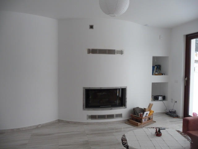 Białe wnętrze salonu z zaokrągloną ścianą, wbudowanym kominkiem z metalowym obramowaniem, dwoma wnękami w ścianie i okrągłym stołem z haftowanym obrusem.