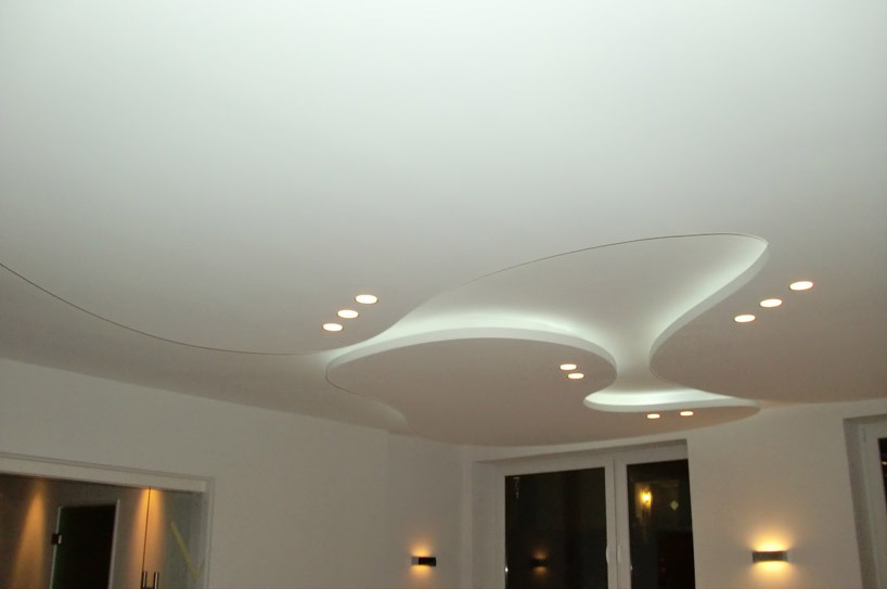 Nowoczesny, biały sufit napinany z nieregularnymi kształtami i wbudowanymi punktami świetlnymi, widok na fragment pomieszczenia z oknami i drzwiami.