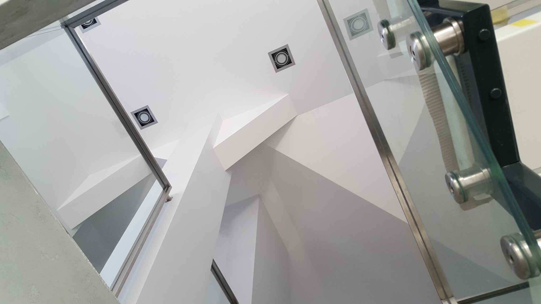 Nowoczesne wnętrze z białymi ścianami, sufit podwieszany z oświetleniem punktowym i szklaną balustradą mocowaną na metalowych uchwytach, widok z dołu.