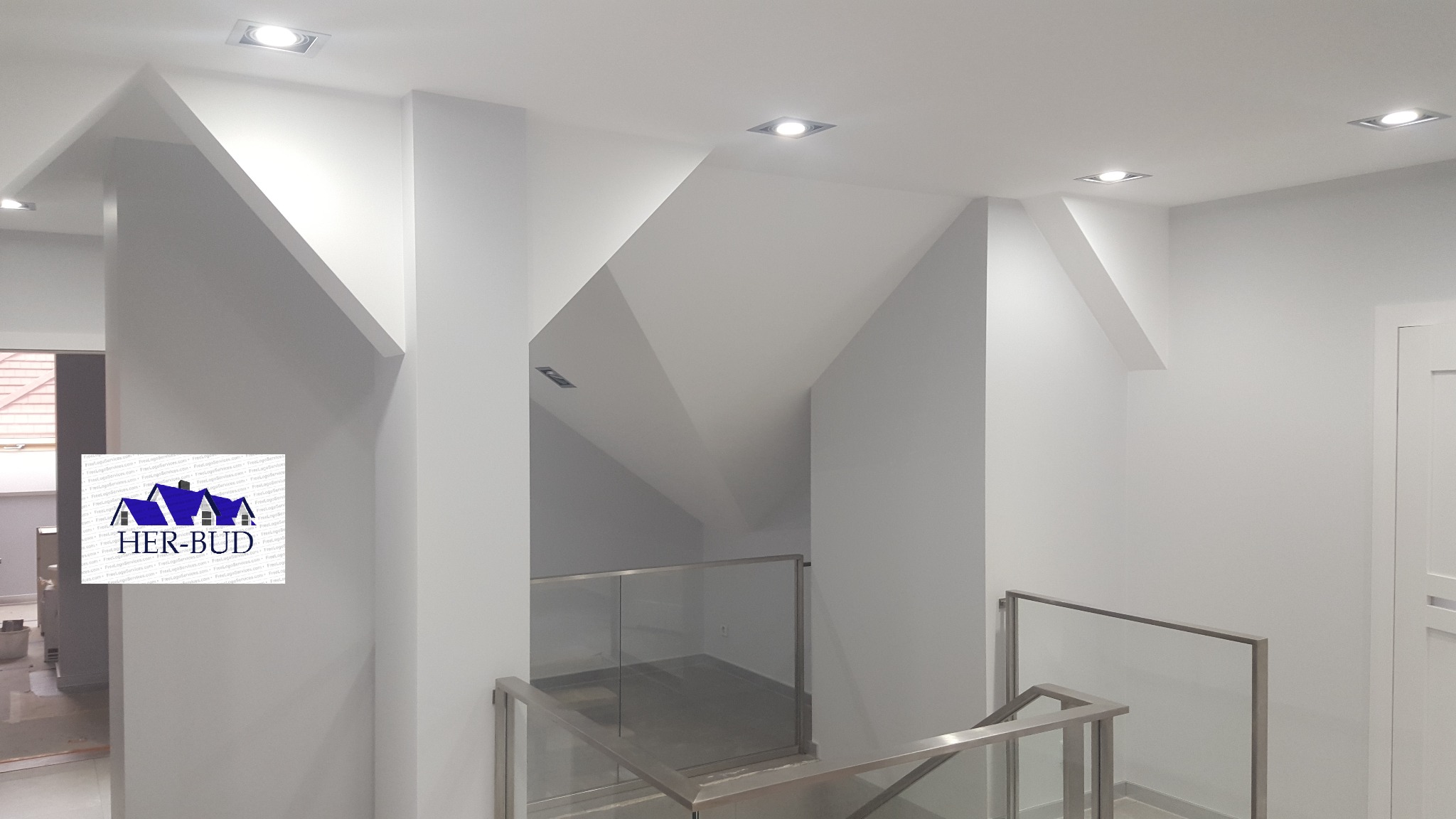 Białe wnętrze z niestandardowym układem sufitu, oświetleniem punktowym i szklaną balustradą schodów, widoczne logo firmy Her-Bud w lewym dolnym rogu.