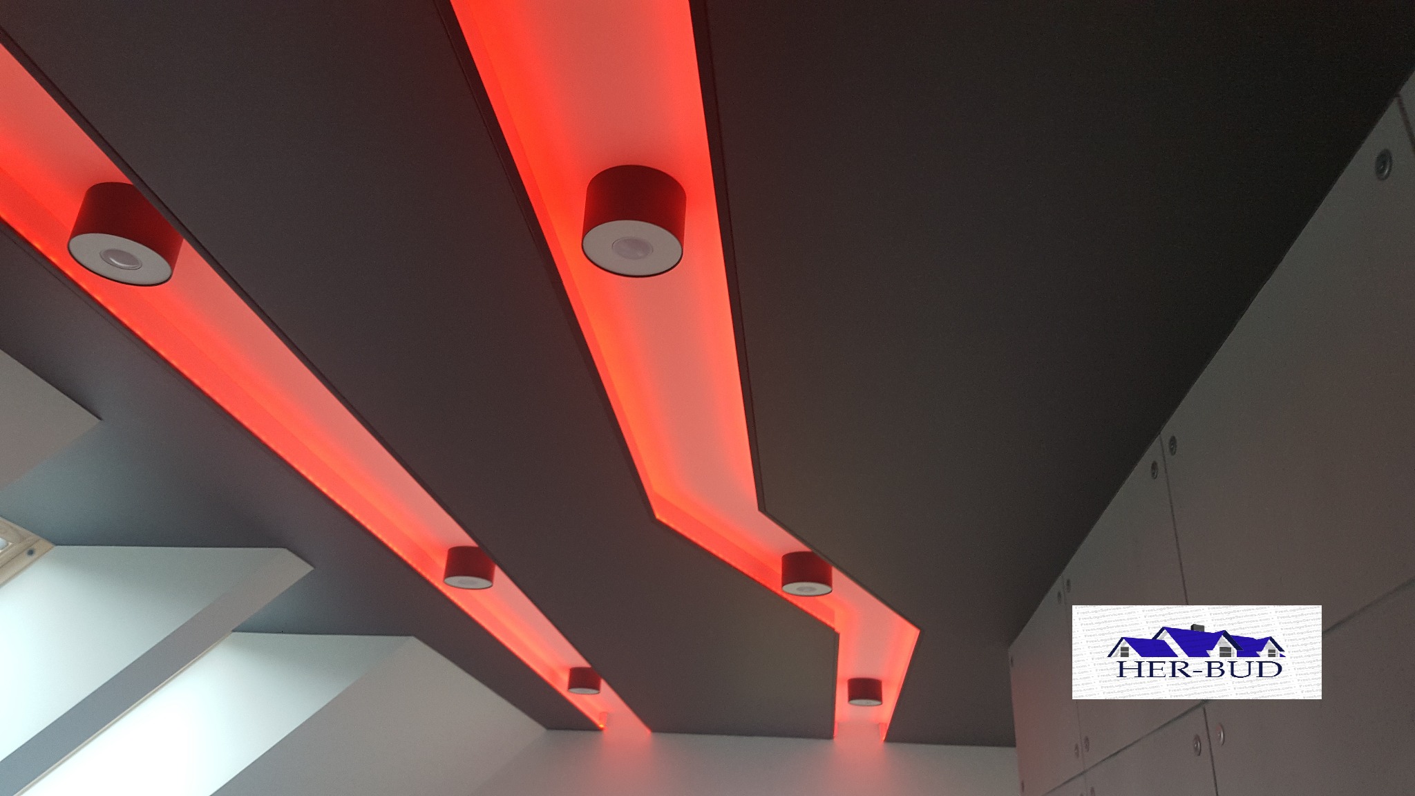 Nowoczesny sufit podwieszany z płyty karton gips, podświetlony czerwonymi lampami LED i okrągłymi spotami, z logo firmy Her-Bud w prawym dolnym rogu.