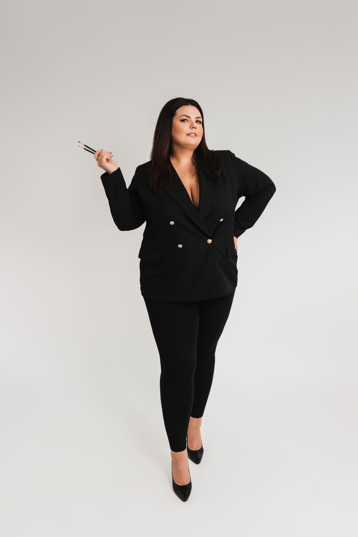 Portret kobiety w czarnym garniturze, trzymającej pędzle do makijażu na jasnym tle, pozującej z ręką na biodrze.