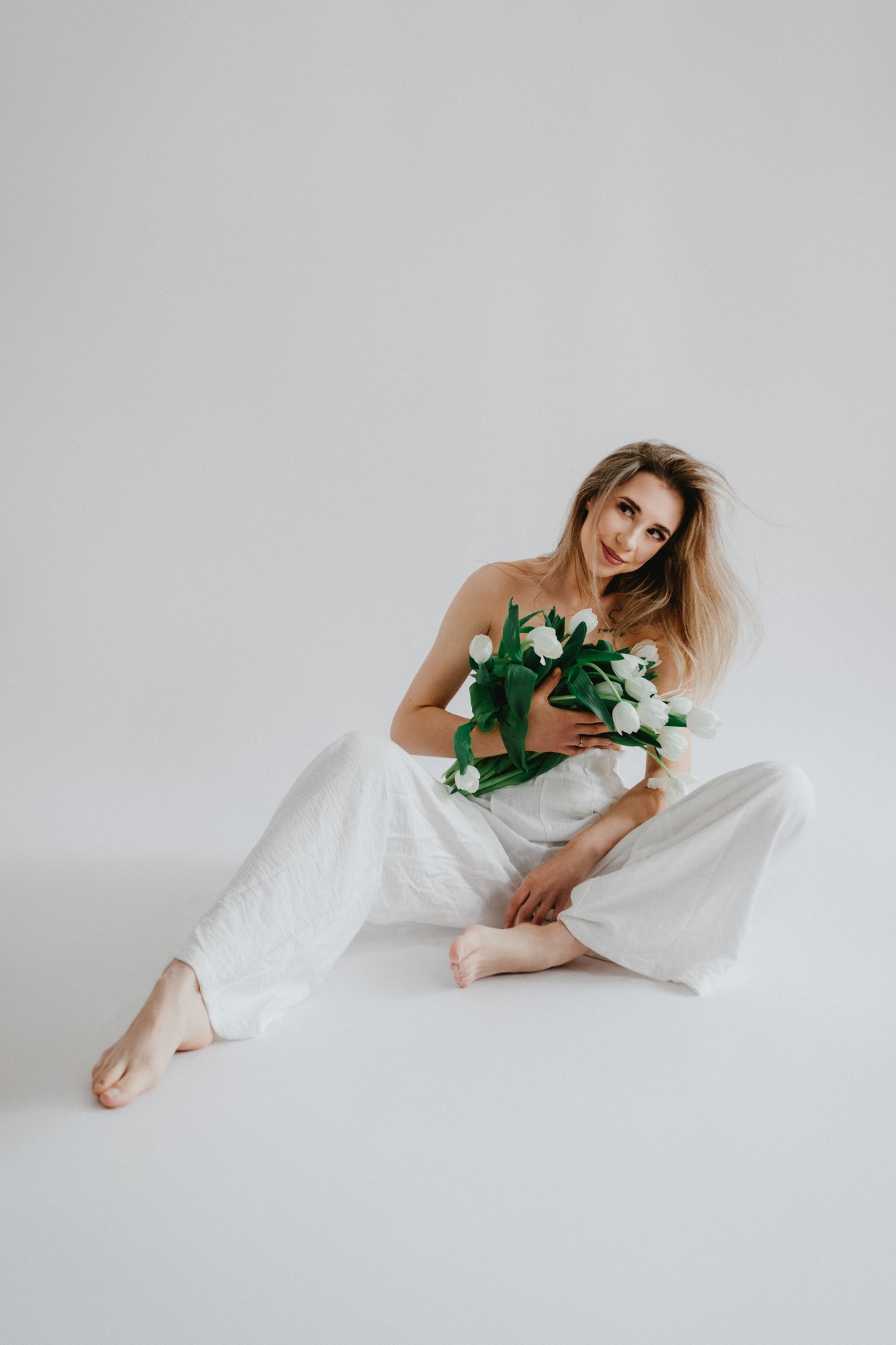 Kobieta w białym kombinezonie siedzi na białym tle, trzymając bukiet białych tulipanów, z delikatnym uśmiechem i rozwianymi włosami.