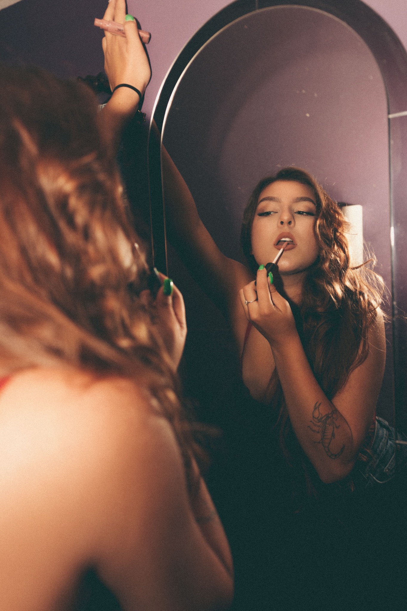 Młoda kobieta z tatuażem skorpiona na ramieniu, aplikuje błyszczyk przed lustrem w łazience z fioletowymi ścianami.