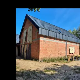 Budynek w trakcie budowy z cegły, z zamontowanym czarnym dachem z blachy na rąbek stojący i oknem dachowym; na ścianach widoczne izolacje w rolkach z nadrukiem producenta.