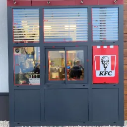 Elewacja okienka drive-thru restauracji KFC z widocznym logo, żaluzjami w oknach i pracownikiem w środku.