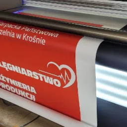 Drukarka wielkoformatowa w trakcie drukowania baneru z logo i nazwą uczelni Karpaciej Państwowej Uczelni w Krośnie oraz hasłem PIELĘGNIARSTWO INŻYNIERIA PRODUKCJI na czerwonym tle.