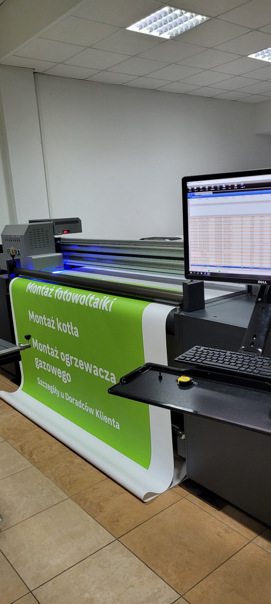 Drukarka wielkoformatowa Flora drukuje zielony baner z białym tekstem: Montaż fotowoltaiki, Montaż kotła, Montaż ogrzewacza gazowego, Szczegóły u Doradców Klienta. Widoczny monitor Dell z danymi...