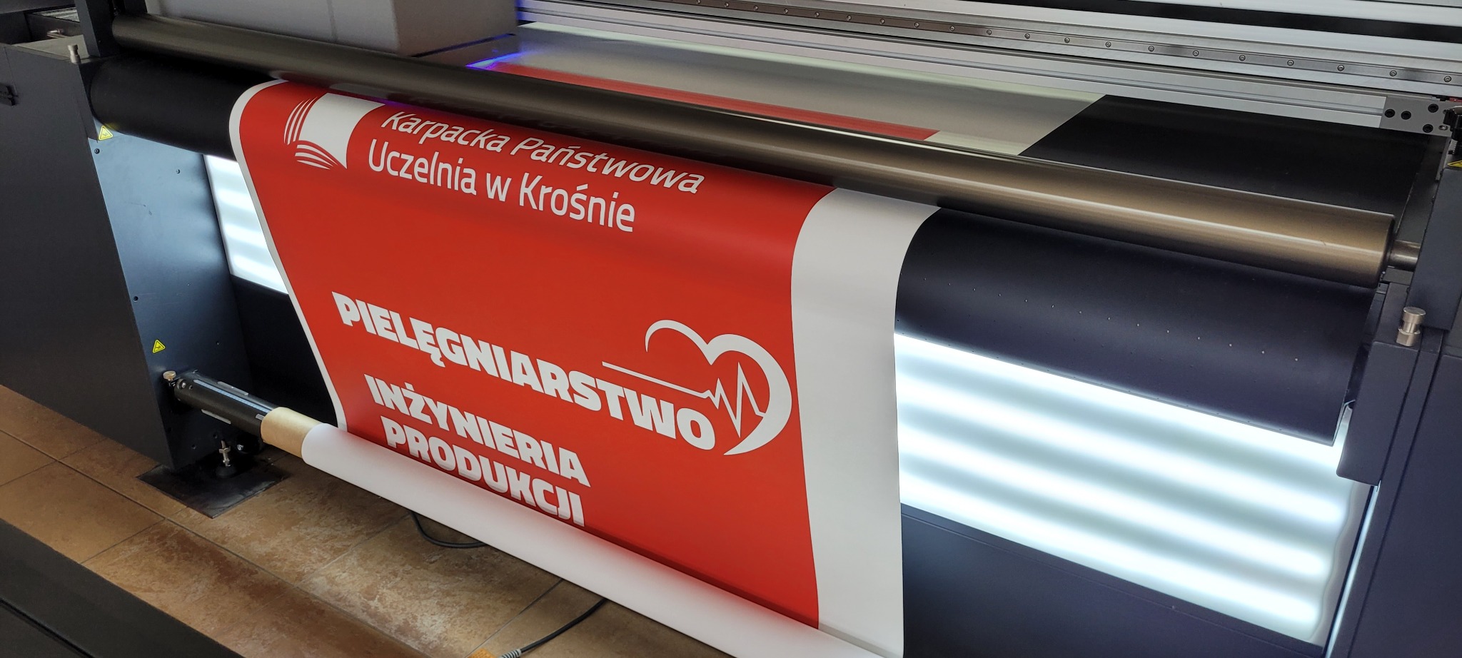 Drukarka wielkoformatowa w trakcie drukowania baneru z logo i nazwą uczelni Karpaciej Państwowej Uczelni w Krośnie oraz hasłem PIELĘGNIARSTWO INŻYNIERIA PRODUKCJI na czerwonym tle.