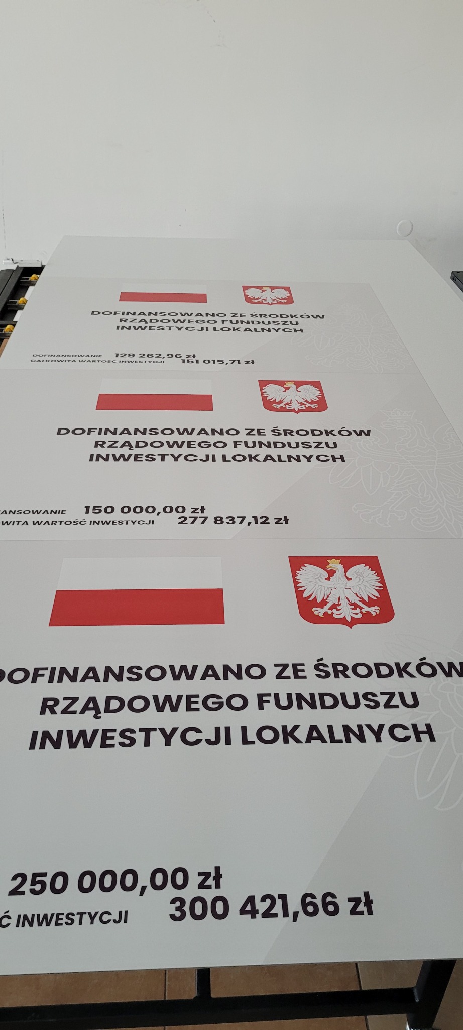 Wielkoformatowy wydruk z informacją o dofinansowaniu z rządowego funduszu inwestycji lokalnych, zawierający flagę i godło Polski oraz kwoty dofinansowania i wartości inwestycji.