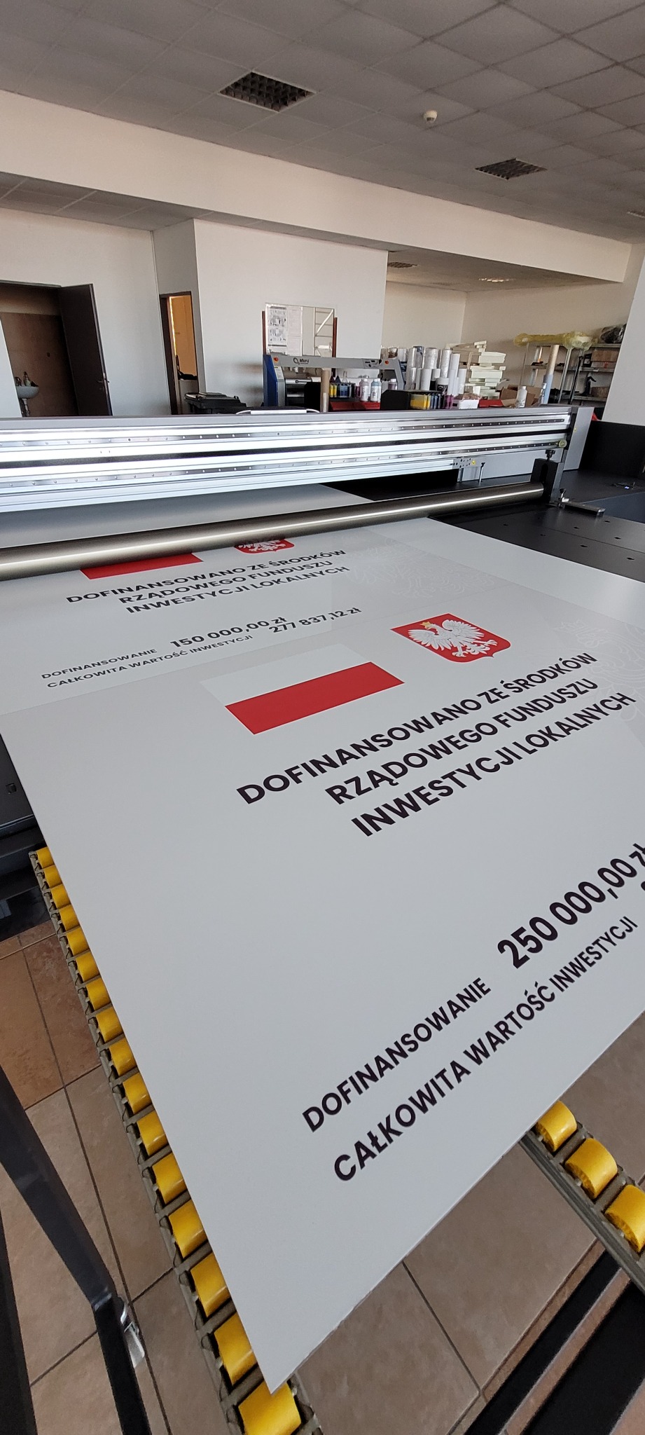 Wielkoformatowy wydruk plakatu informacyjnego z flagą Polski, na stole drukarki przemysłowej, w tle widoczny sprzęt drukarski i materiały eksploatacyjne.