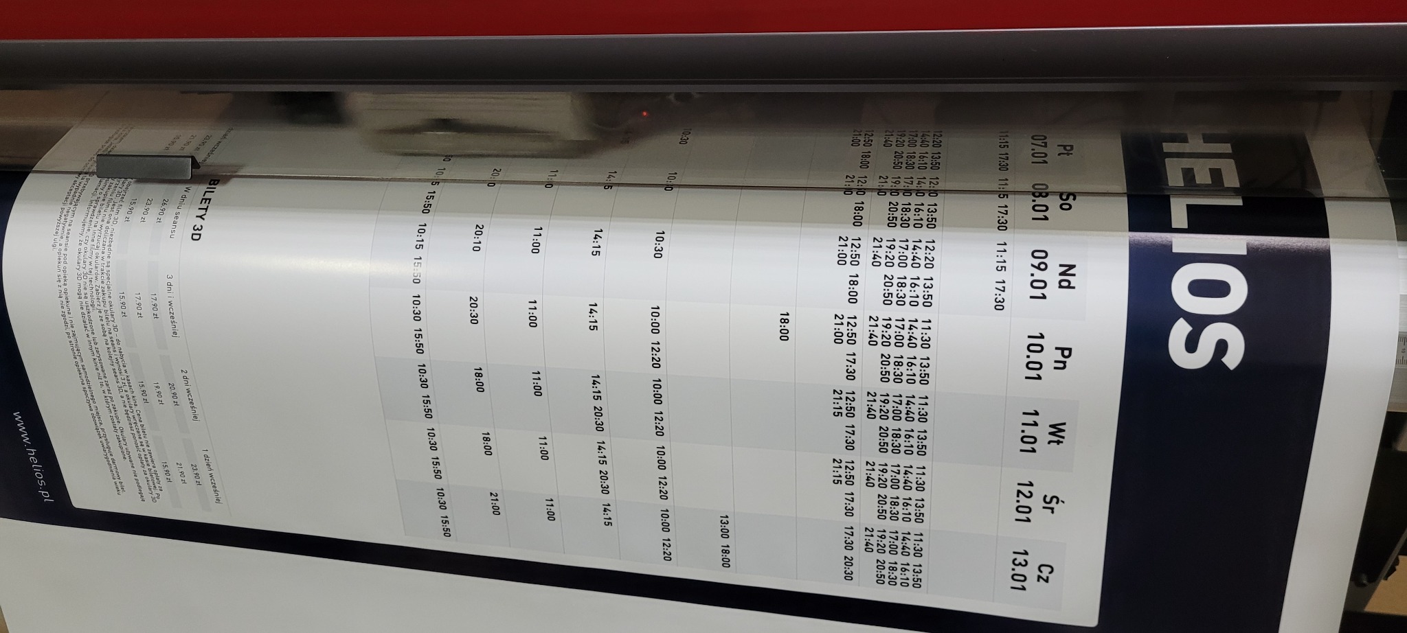 Wielkoformatowy wydruk plakatu z repertuarem kina Helios, ukazujący dni tygodnia i godziny seansów 3D, w trakcie procesu drukowania.