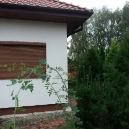 Brązowa osłona okienna zamontowana na elewacji domu z czerwonym dachem, widok z poziomu ogrodu z krzewami na pierwszym planie.