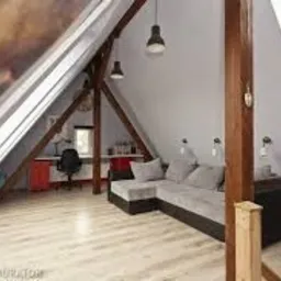 Adaptacja poddasza: skośne ściany pomalowane na biało, drewniane belki konstrukcyjne, szara narożna sofa, biurko z czerwonym kontenerkiem pod oknem dachowym, jasna podłoga z paneli.