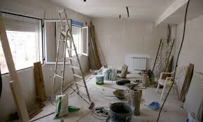 Pokój w trakcie remontu: widoczne ślady po szpachlowaniu na ścianach, drabina oparta o okno, worki z materiałami budowlanymi, narzędzia malarskie i resztki gruzu na podłodze. Oświetlenie górne...