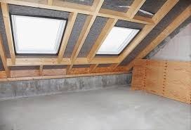 Surowe poddasze ze skośnym sufitem, widoczne drewniane belki konstrukcyjne, dwa okna dachowe, szara izolacja i surowa betonowa podłoga. Część ściany wykończona płytą OSB.