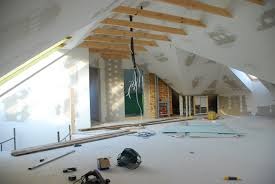 Poddasze w trakcie prac wykończeniowych: surowe ściany, widoczne elementy konstrukcji drewnianej dachu, płyty gipsowo-kartonowe na podłodze, narzędzia budowlane w nieładzie.