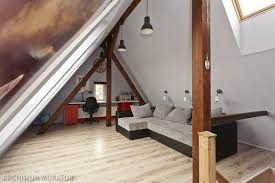 Adaptacja poddasza: skośne ściany pomalowane na biało, drewniane belki konstrukcyjne, szara narożna sofa, biurko z czerwonym kontenerkiem pod oknem dachowym, jasna podłoga z paneli.