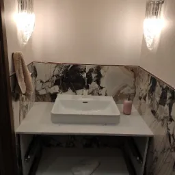 Nowoczesna umywalka nablatowa o kwadratowym kształcie, osadzona na marmurowym blacie z otwartą szafką, oświetlona dwoma szklanymi kinkietami, z marmurowymi ścianami w tle.