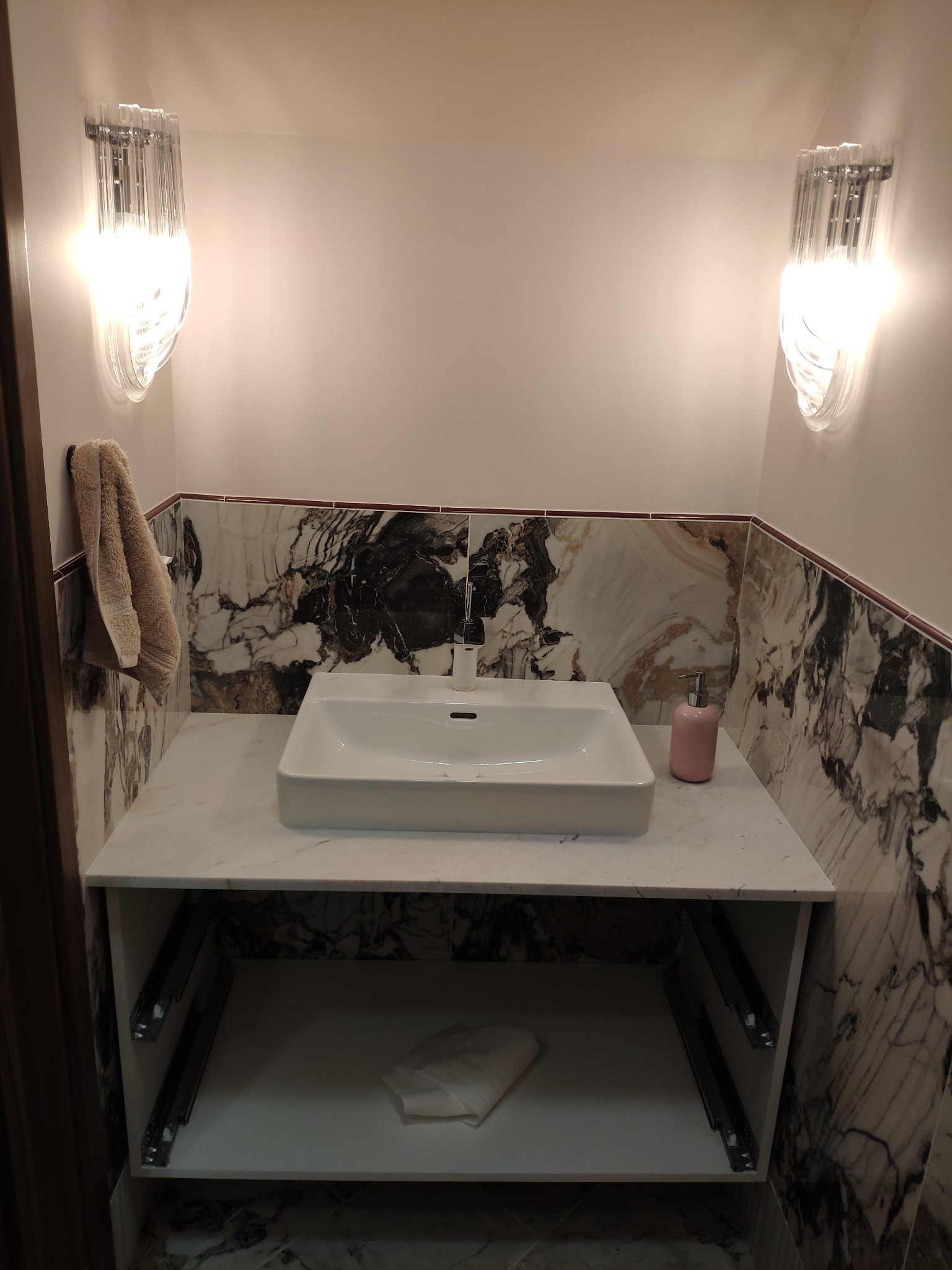 Nowoczesna umywalka nablatowa o kwadratowym kształcie, osadzona na marmurowym blacie z otwartą szafką, oświetlona dwoma szklanymi kinkietami, z marmurowymi ścianami w tle.