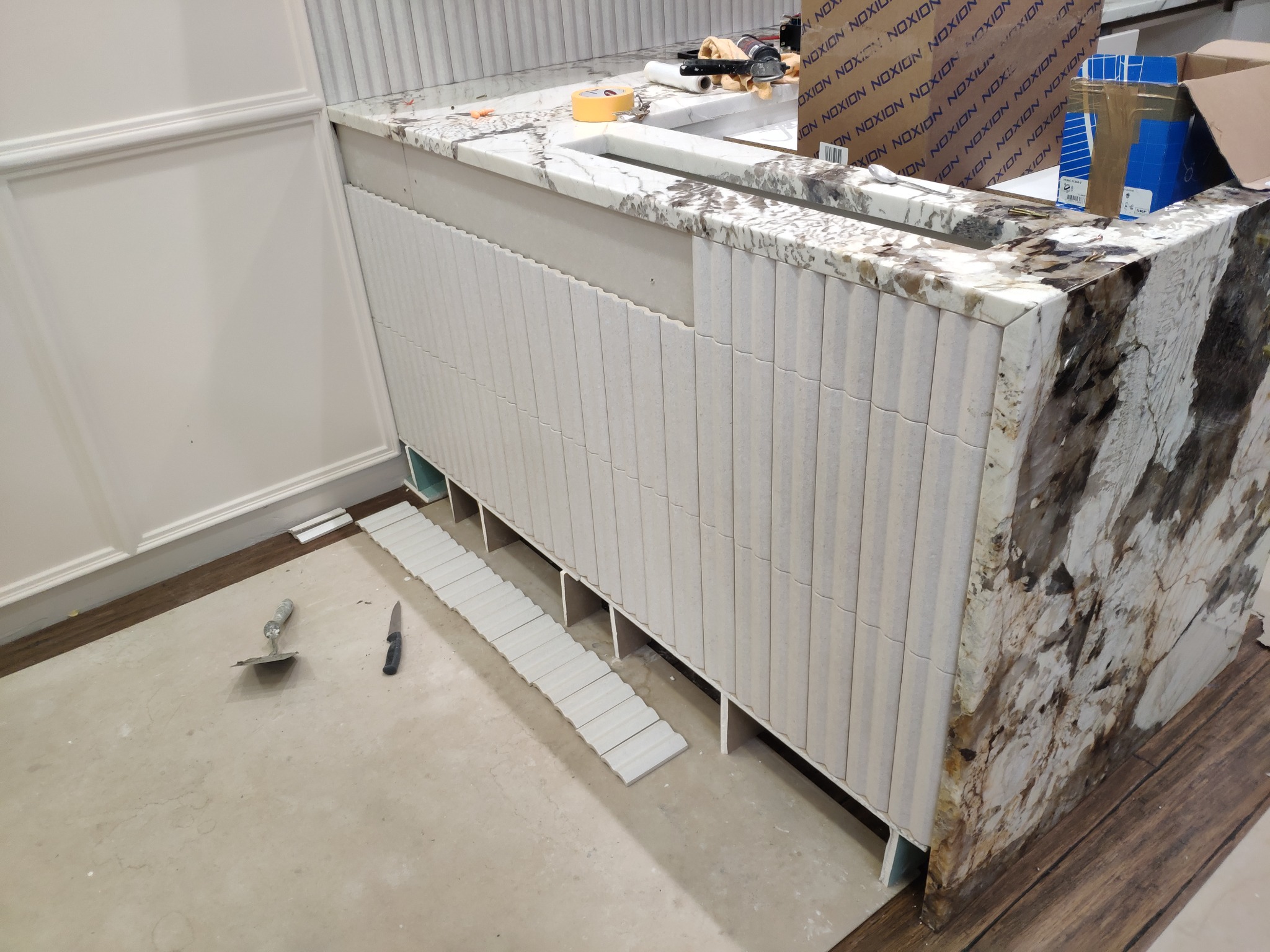 Montaż dekoracyjnych, pionowych płytek na obudowie wanny lub umywalki z blatem z kamienia naturalnego, widoczne narzędzia i kartony w tle.