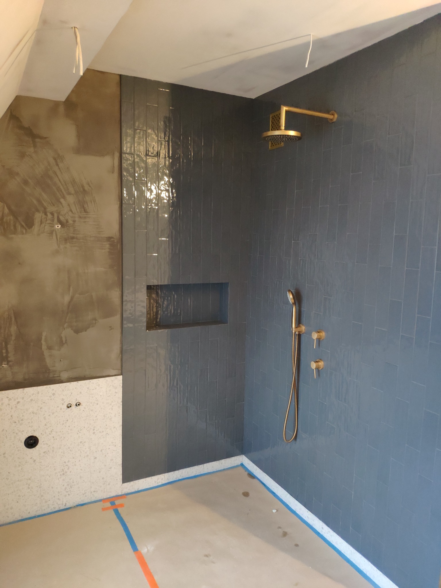 Wnętrze łazienki w Kobiórze w trakcie prac wykończeniowych, widoczne szare, pionowo ułożone płytki, złota armatura, wnęka w ścianie, fragment ściany z szpachlą i fragment ściany z białymi płytkami...