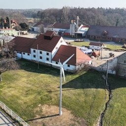 ISATO POLSKA SPÓŁKA Z OGRANICZONĄ ODPOWIEDZIALNOŚCIĄ - Widok z góry na farmę z turbiną wiatrową na pierwszym planie, panele fotowoltaiczne widoczne na dachu budynku po lewej stronie, w tle zabudowania gospodarcze i drzewa.