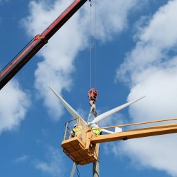 ISATO POLSKA SPÓŁKA Z OGRANICZONĄ ODPOWIEDZIALNOŚCIĄ - Montaż łopat turbiny wiatrowej na maszcie przy użyciu dźwigu i podnośnika koszowego na tle błękitnego nieba z chmurami.