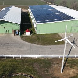 ISATO POLSKA SPÓŁKA Z OGRANICZONĄ ODPOWIEDZIALNOŚCIĄ - Widok z góry na dwa zielone budynki z panelami słonecznymi na dachu i turbiną wiatrową na pierwszym planie, białe ogrodzenie, dwa samochody zaparkowane obok siebie.