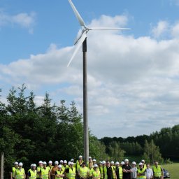 ISATO POLSKA SPÓŁKA Z OGRANICZONĄ ODPOWIEDZIALNOŚCIĄ - Grupa osób w odblaskowych kamizelkach i kaskach pozuje na tle turbiny wiatrowej przy słonecznej pogodzie.