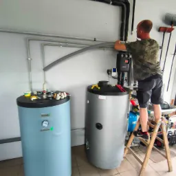 Instalator na drabinie łączy rury do zasobnika ciepłej wody użytkowej w kotłowni; widoczne dodatkowe narzędzia i elementy instalacji.