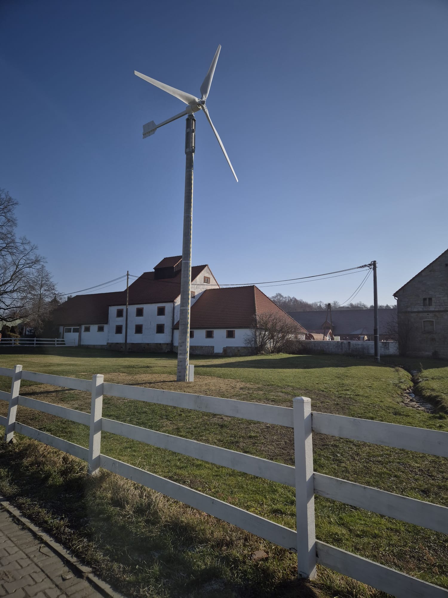 Mała turbina wiatrowa z trzema łopatami na tle wiejskiego krajobrazu z białym płotem i budynkami gospodarczymi.