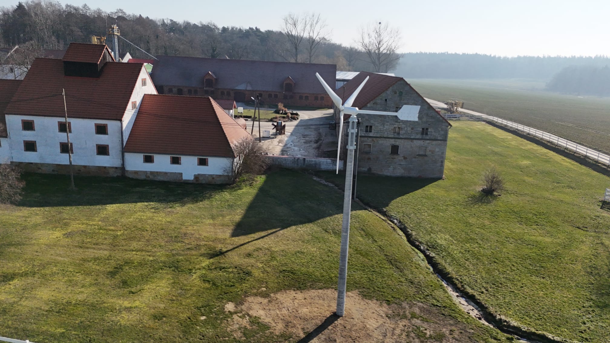 Mała turbina wiatrowa z trzema śmigłami zainstalowana na zielonej łące obok zabudowań gospodarczych w słoneczny dzień.