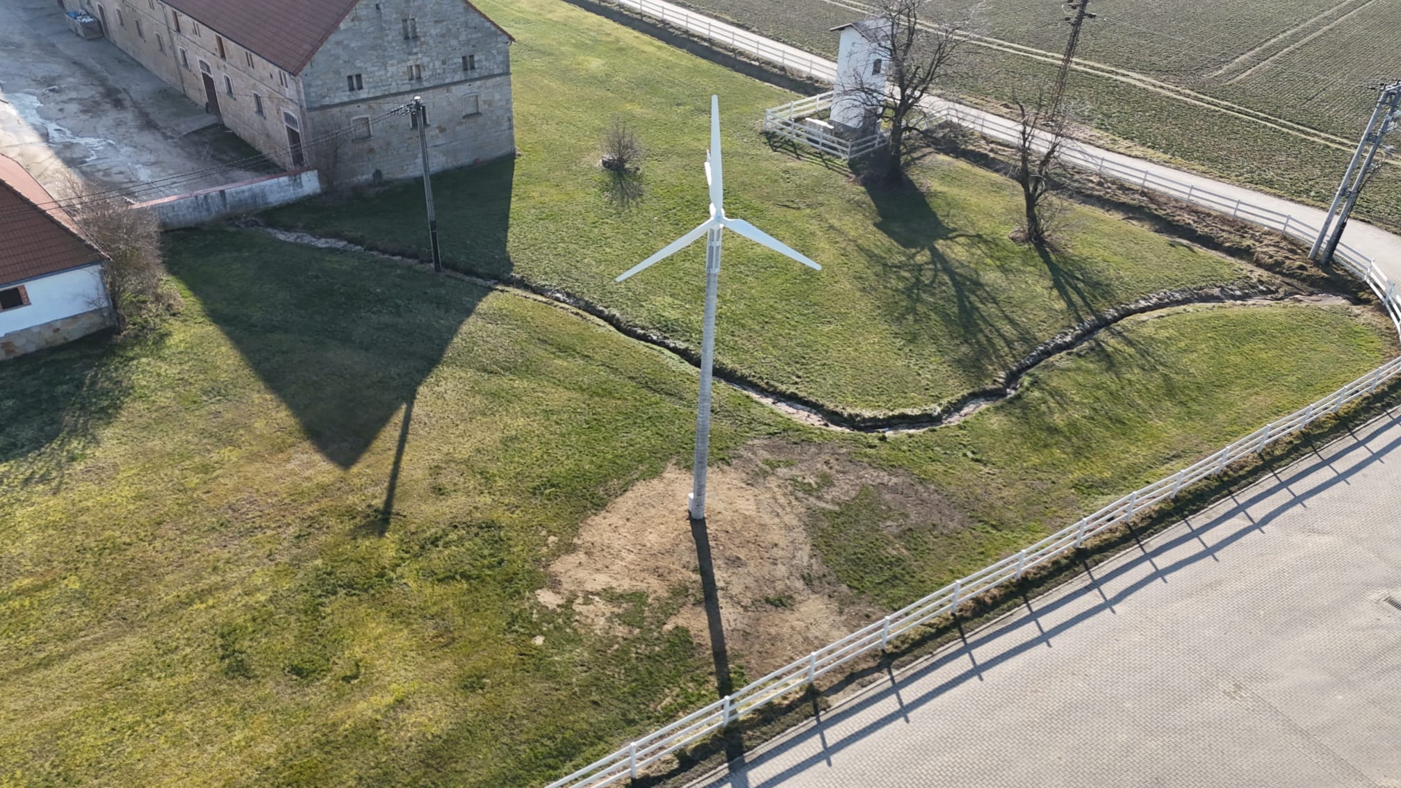 Widok z góry na małą turbinę wiatrową z trzema śmigłami na zielonym terenie wiejskim, obok budynki gospodarcze i pole uprawne.