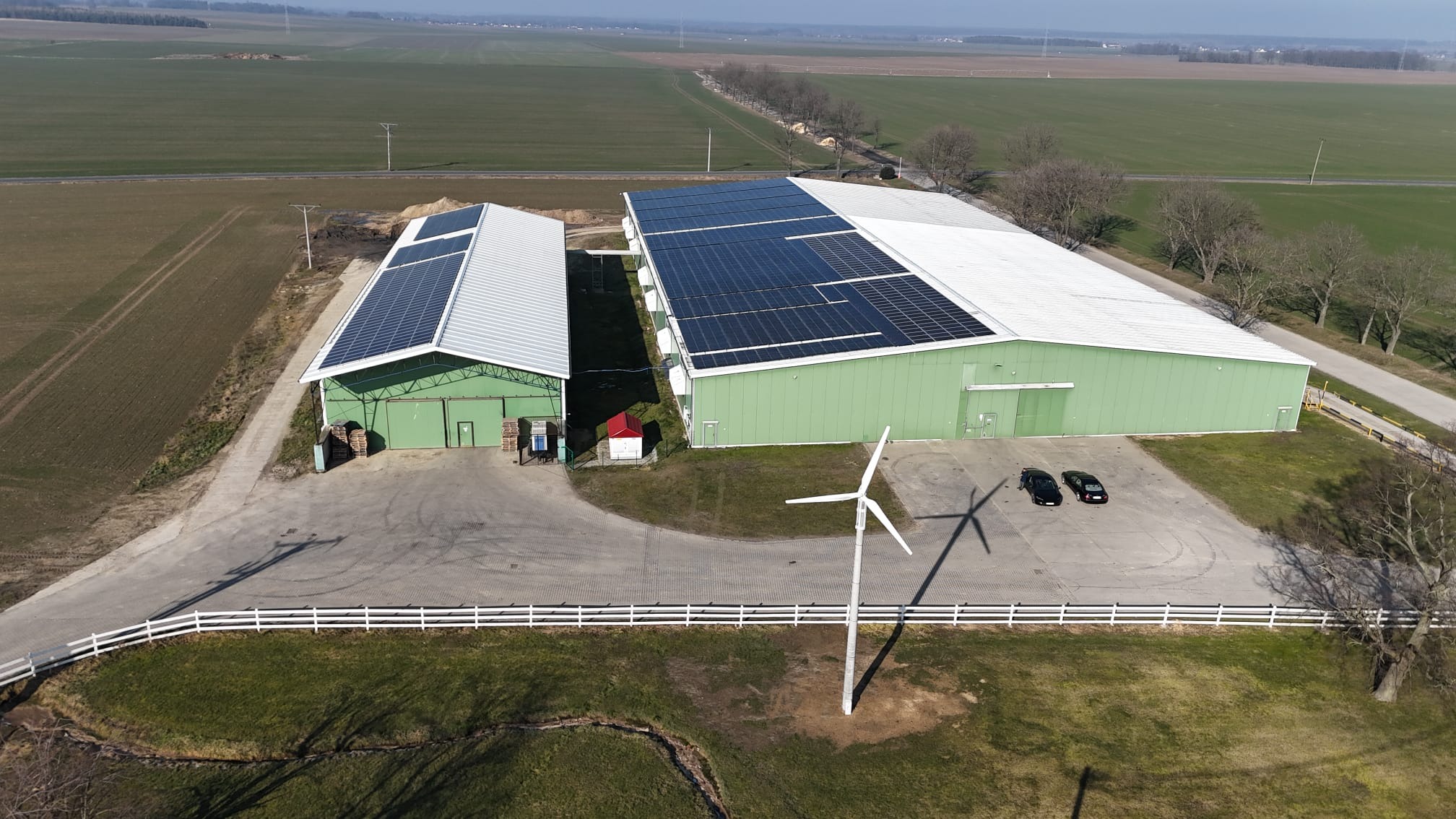 Widok z lotu ptaka na dwa zielone budynki z panelami słonecznymi na dachu, turbina wiatrowa na pierwszym planie, dwa samochody zaparkowane obok większego budynku, pola uprawne w tle.