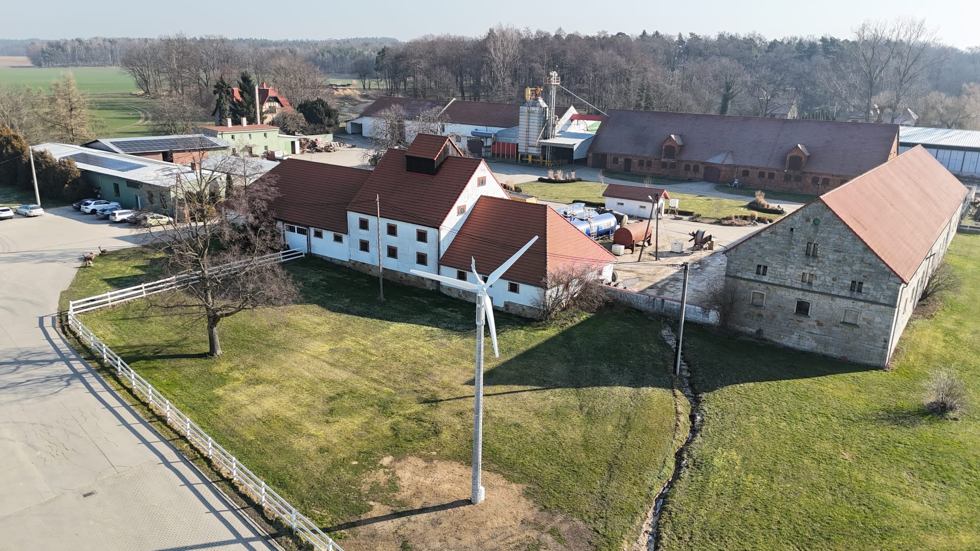 Widok z góry na farmę z turbiną wiatrową na pierwszym planie, panele fotowoltaiczne widoczne na dachu budynku po lewej stronie, w tle zabudowania gospodarcze i drzewa.
