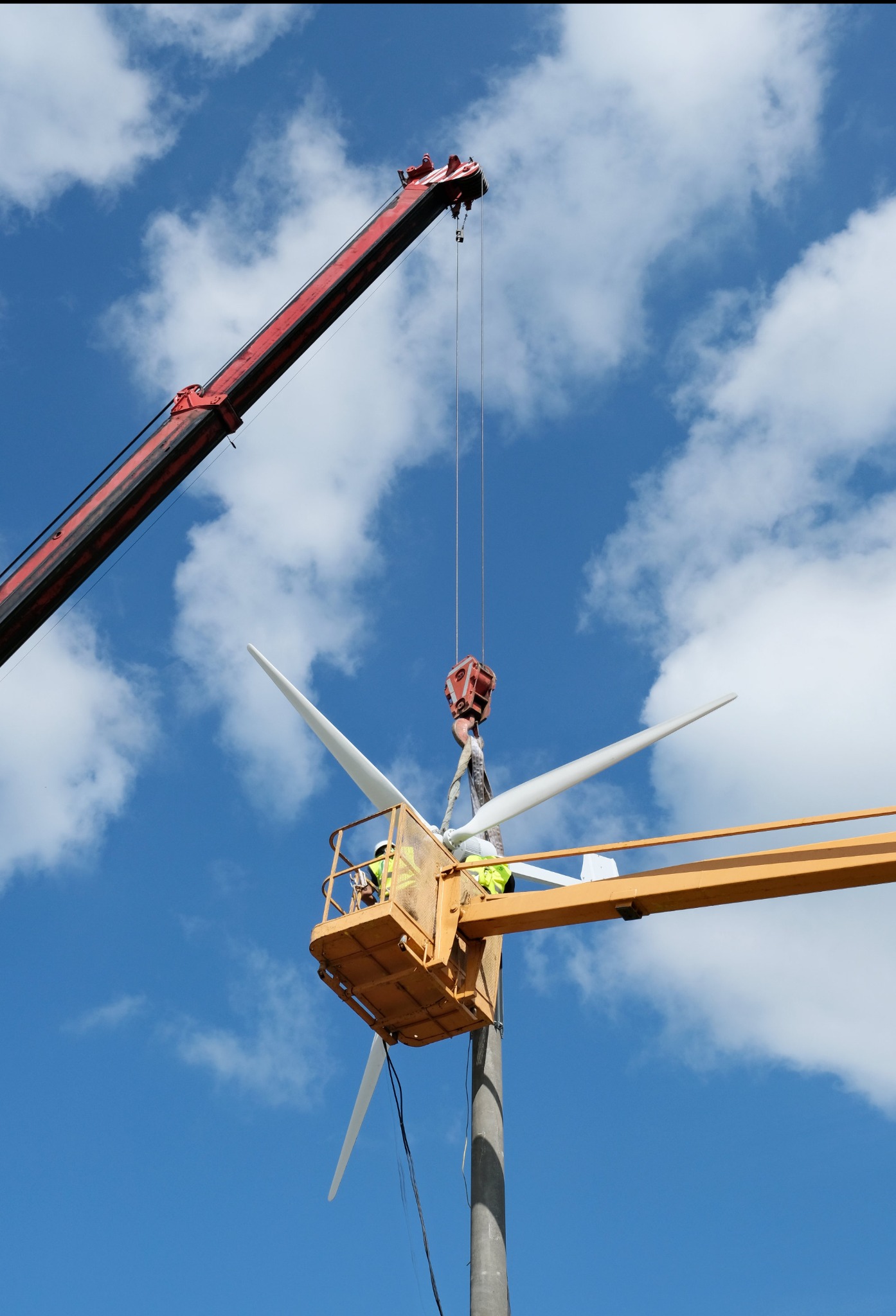 Montaż łopat turbiny wiatrowej na maszcie przy użyciu dźwigu i podnośnika koszowego na tle błękitnego nieba z chmurami.