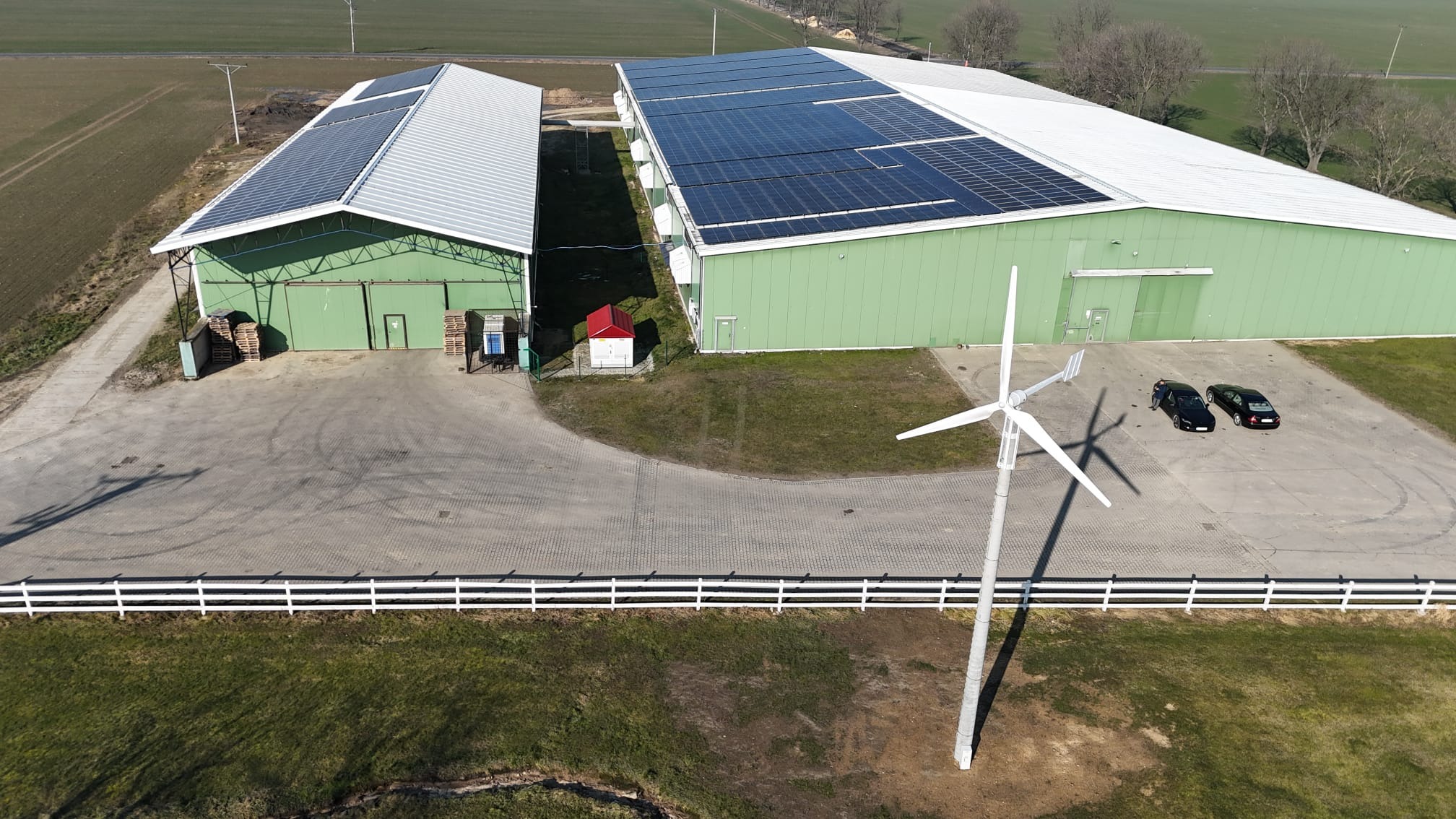 Widok z góry na dwa zielone budynki z panelami słonecznymi na dachu i turbiną wiatrową na pierwszym planie, białe ogrodzenie, dwa samochody zaparkowane obok siebie.