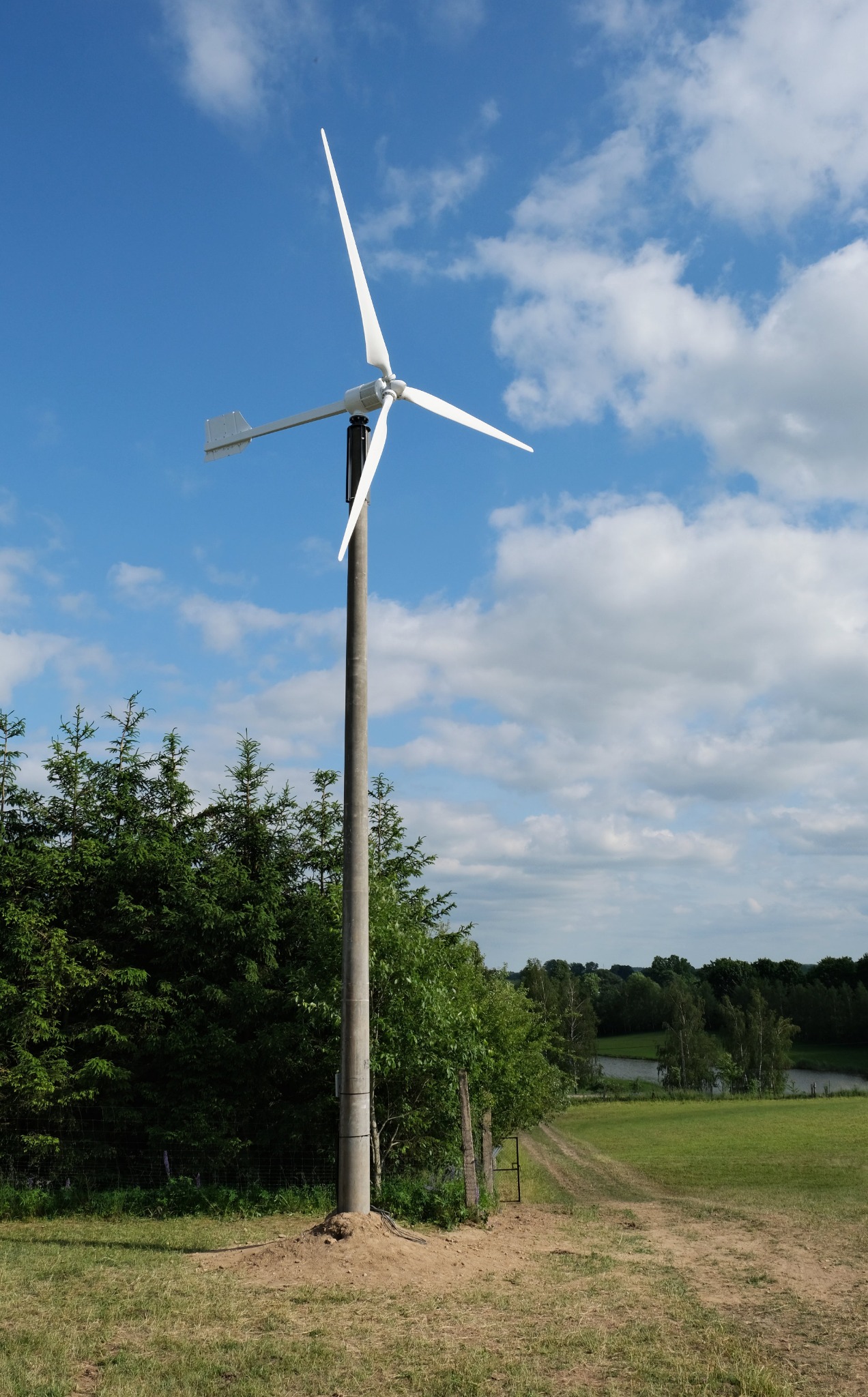 Niewielka turbina wiatrowa na betonowym słupie w Łodzi, z trzema białymi śmigłami na tle błękitnego nieba z chmurami, otoczona zielenią drzew i trawy, z widoczną ścieżką polną w tle.
